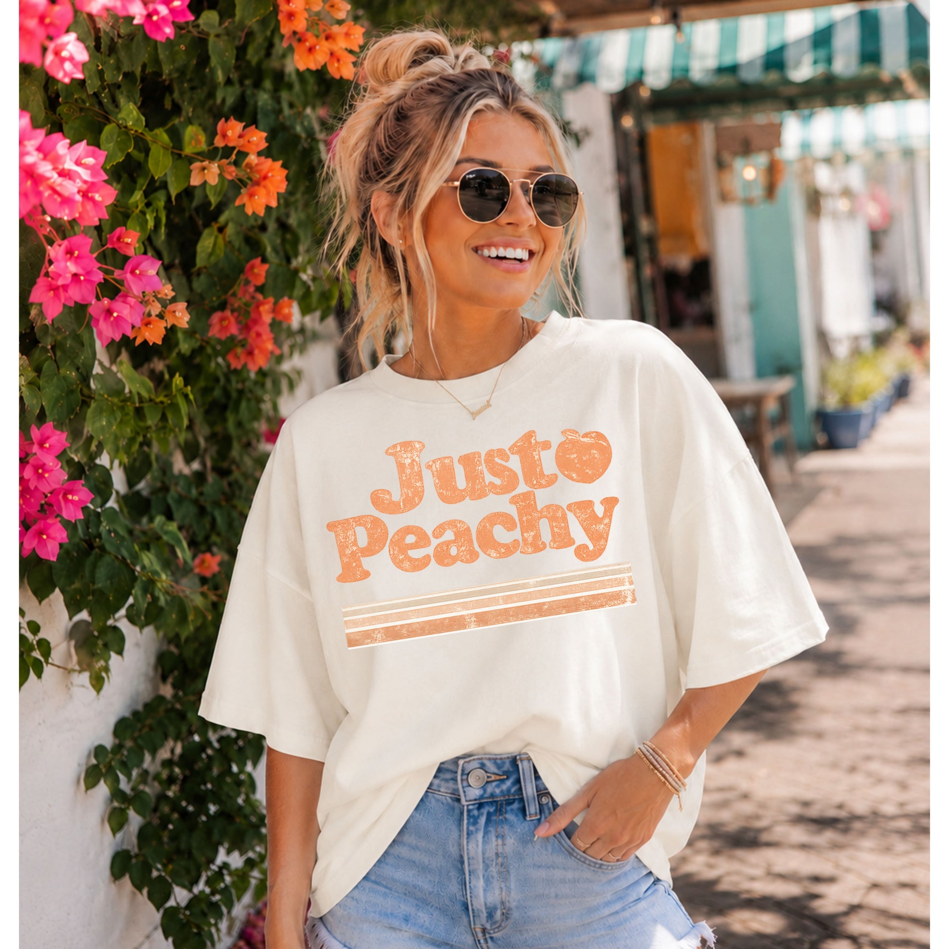 Smiling woman wearing an oversized cream "Just Peachy" graphic T-shirt with a vintage distressed peach design, styled with light wash denim and sunglasses while walking through a sunny coastal shopping district lined with vibrant bougainvillea.