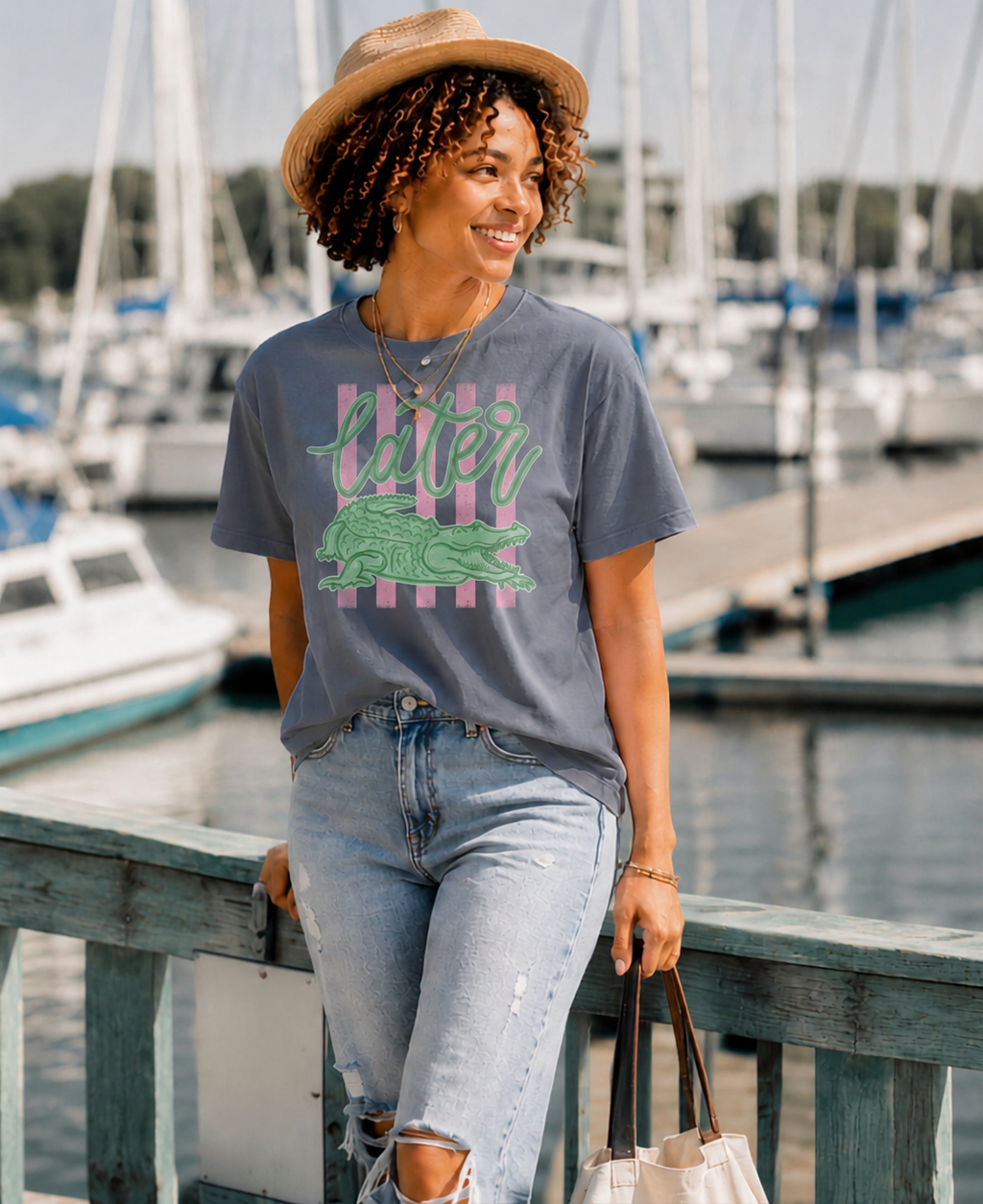 Woman wearing a blue-gray "Later Gator" graphic T-shirt featuring a vintage alligator design while strolling along a sunny marina with sailboats, styled with light-wash denim, a straw hat, and a canvas tote.