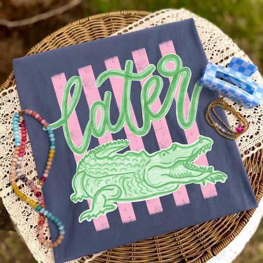 Folded blue-gray graphic T-shirt featuring the "Later Gator" alligator design displayed on a woven tray with colorful beaded bracelets, delicate jewelry, and a blue checkered hair clip.