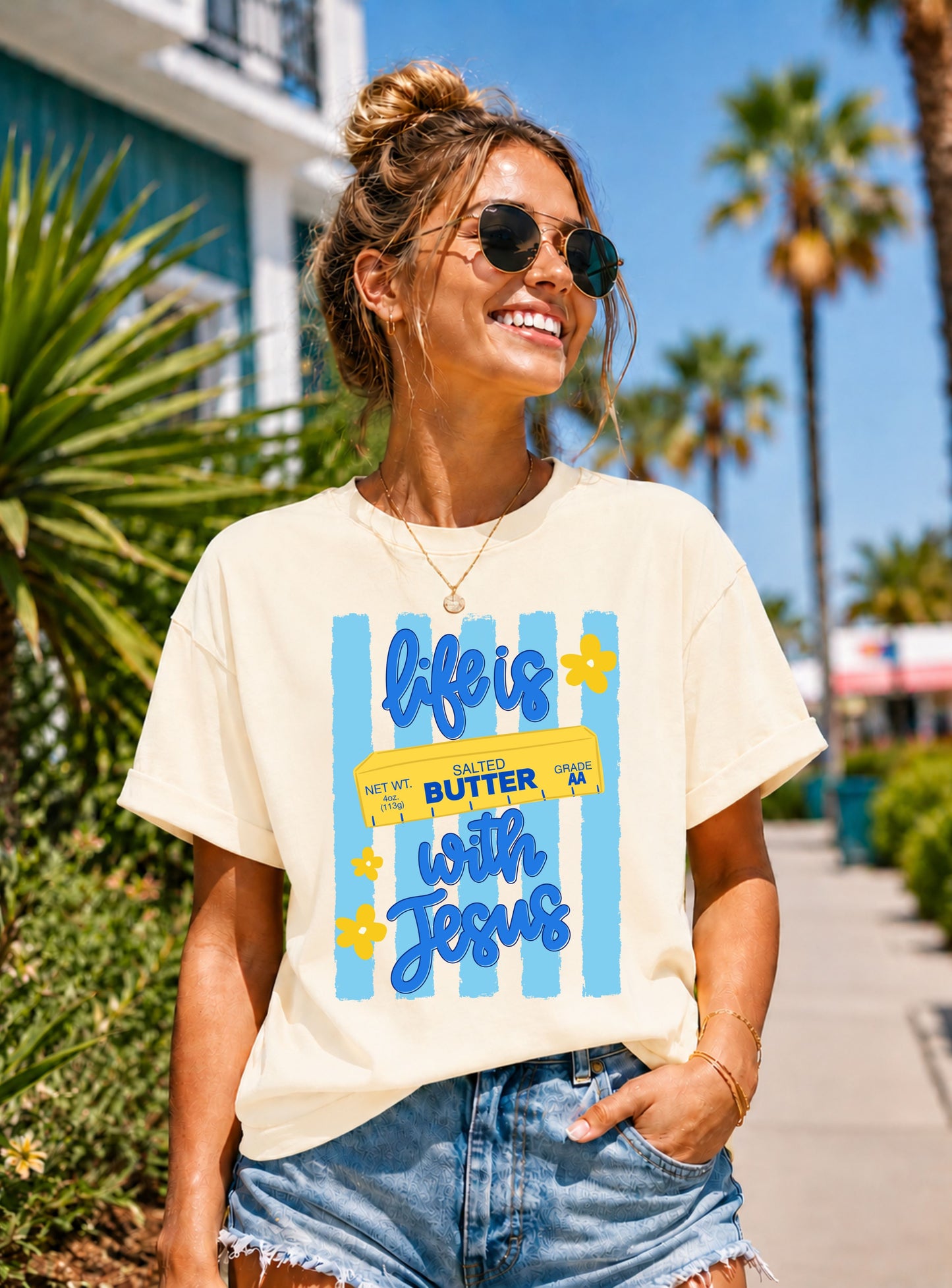 Smiling woman wearing a cream "Life Is Butter With Jesus" graphic T-shirt featuring blue stripes, yellow florals, and a playful butter design while walking along a sunny palm-lined coastal street