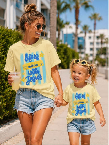 Mom and daughter wearing matching "Life Is Butter With Jesus" graphic T-shirts while walking hand in hand on a sunny palm-lined sidewalk, creating a cheerful faith-filled Mommy & Me summer look.