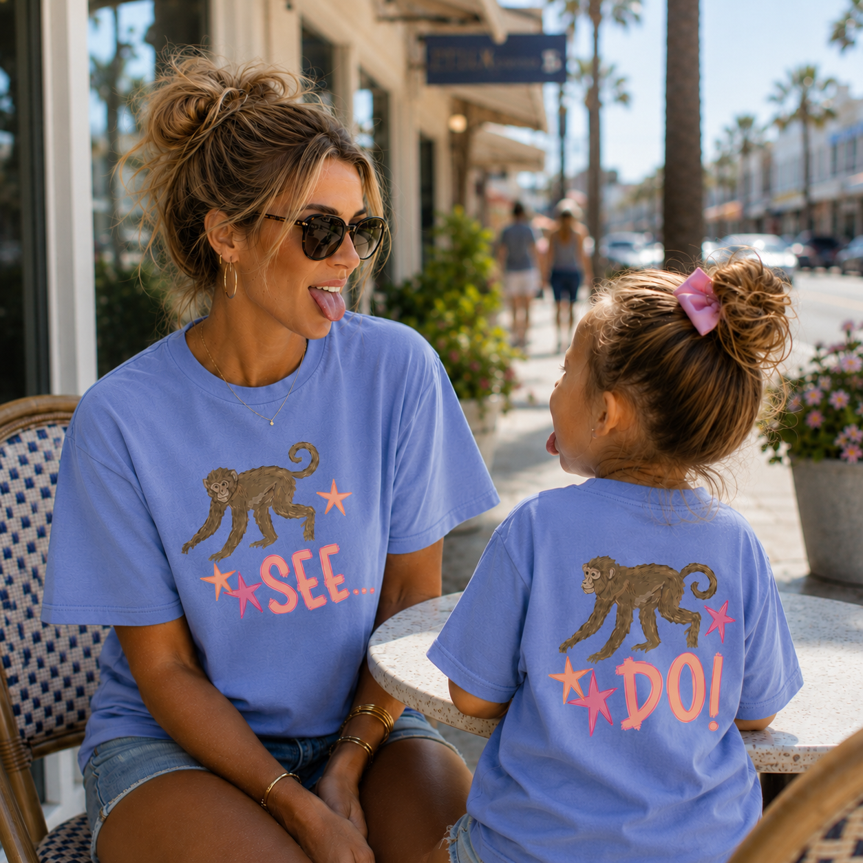 Mom and daughter wearing matching blue "Monkey See, Monkey Do" graphic T-shirts while playfully sticking out their tongues at each other at an outdoor café, showcasing the front "Monkey See..." design on Mom and the back "Monkey Do!" design on the child.