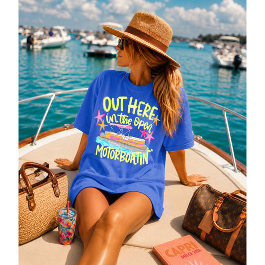 Woman relaxing on the bow of a boat wearing a bright blue "Out Here in the Open Motorboatin'" graphic T-shirt with a colorful pontoon boat design, styled with a straw hat while enjoying a sunny day on the water.