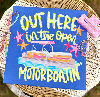 Folded bright blue graphic T-shirt featuring the colorful "Out Here in the Open Motorboatin'" pontoon boat design, styled on a woven tray with pastel beaded bracelets, layered jewelry, and a pink checkered hair clip.