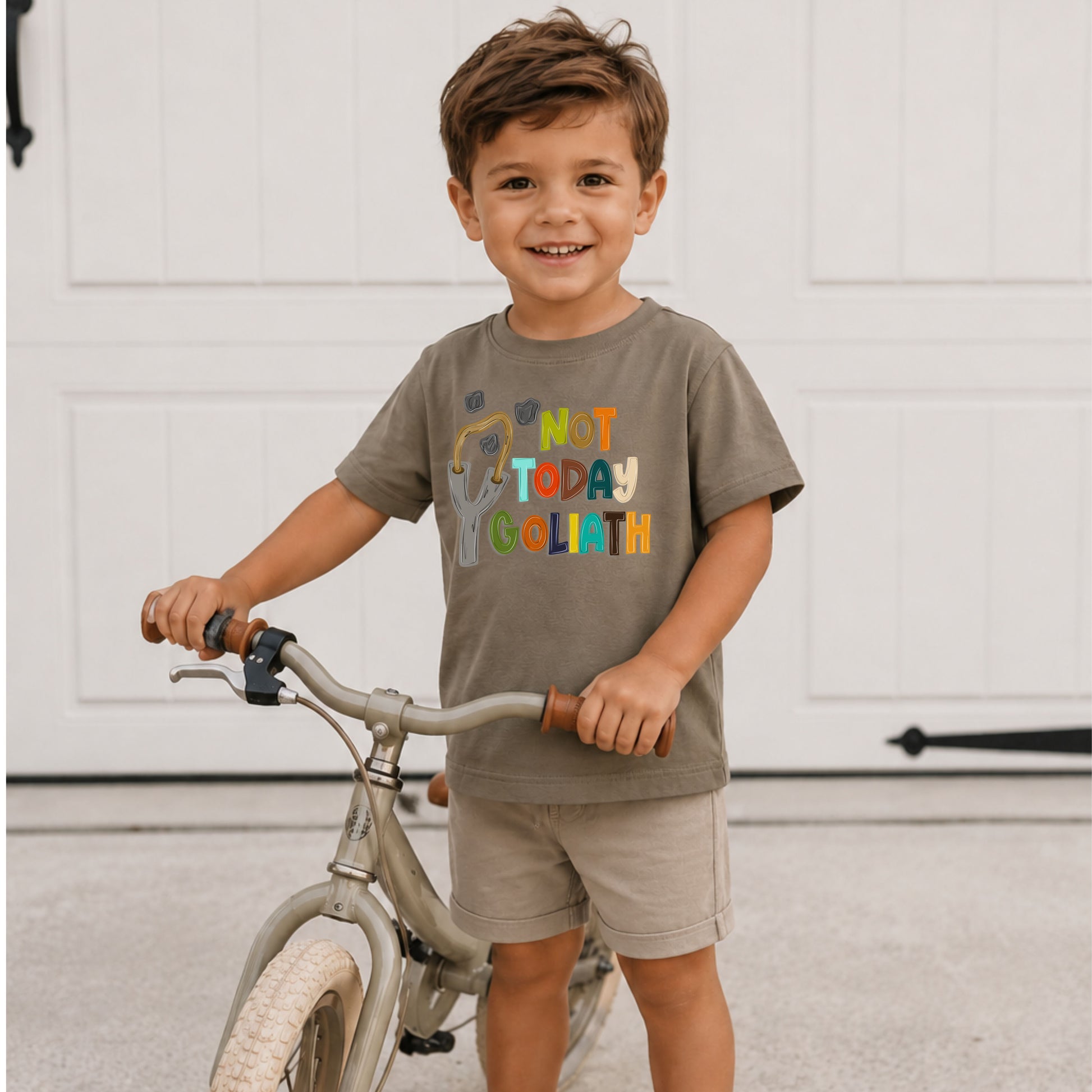 Child wearing a shirt with colorful text standing next to a bicycle indoors.