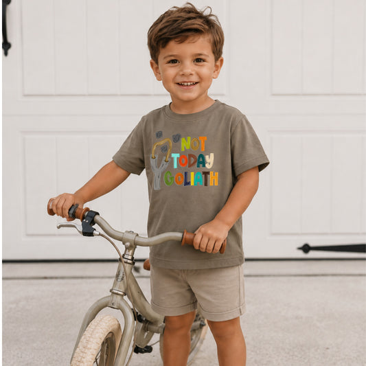 Child wearing a shirt with colorful text standing next to a bicycle indoors.