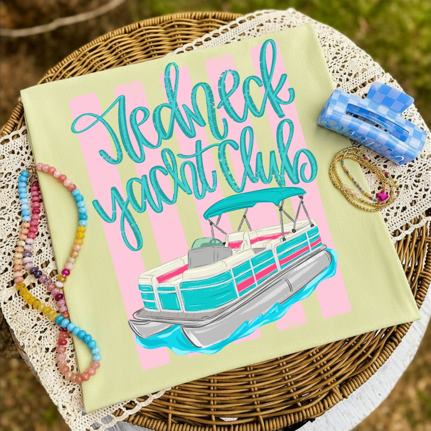Folded butter yellow graphic T-shirt featuring the colorful "Redneck Yacht Club" pontoon boat design, styled on a woven tray with pastel beaded bracelets, layered jewelry, and a blue checkered hair clip.