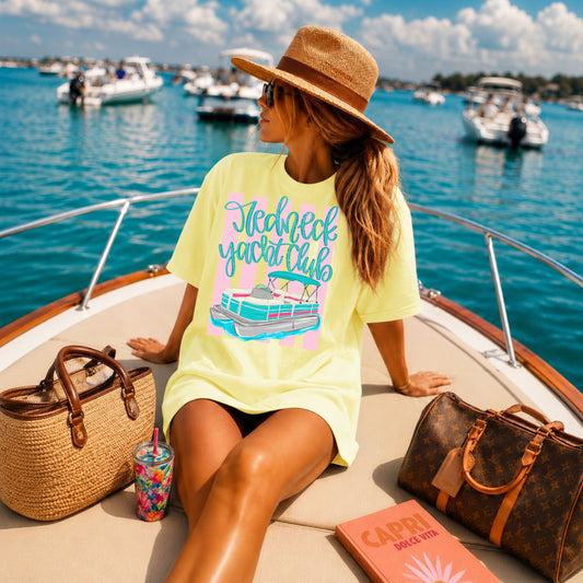Woman relaxing on the bow of a boat wearing a butter yellow "Redneck Yacht Club" graphic T-shirt with a colorful pontoon boat design, styled with a straw hat while enjoying a sunny day on the water.