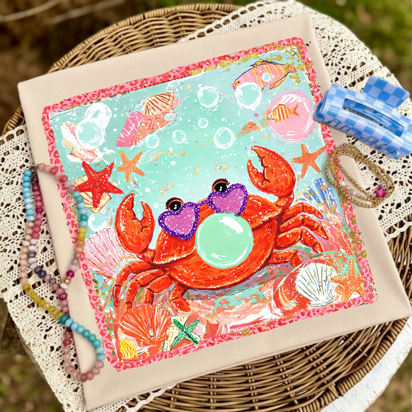 Flat lay of a cream graphic t-shirt featuring a colorful bubblegum-blowing crab surrounded by seashells, starfish, bubbles, and ocean-inspired artwork, styled with pastel beaded bracelets, gold bracelets, and a blue gingham hair clip on a wicker table with lace fabric.