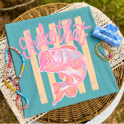 Flat lay of a teal coastal graphic t-shirt featuring a pink fish illustration with vintage cream stripes and the phrase "Shake That," styled with colorful beaded bracelets, gold bracelets, and a blue gingham hair clip on a wicker table with lace fabric.