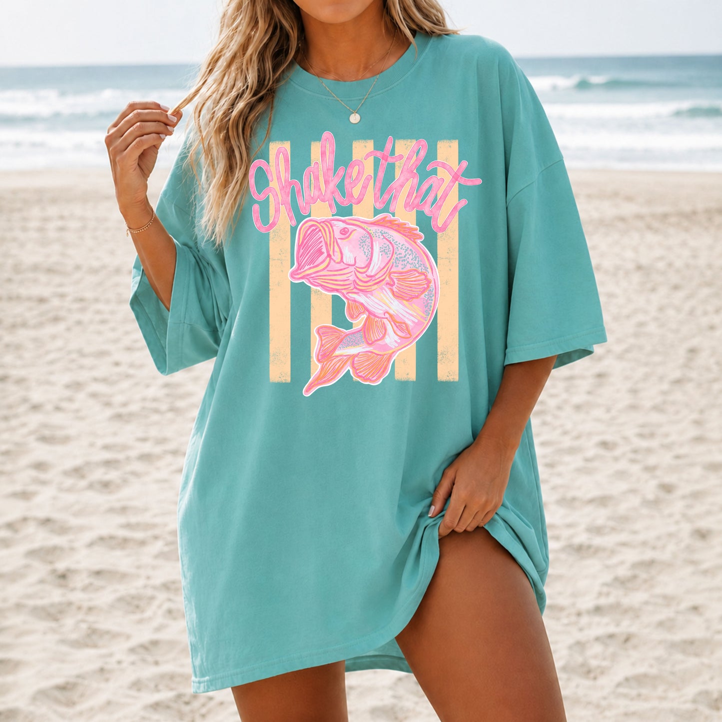 Woman wearing an oversized teal coastal graphic t-shirt featuring a pink fish illustration and the phrase "Shake That" while standing on a sandy beach with ocean waves in the background.