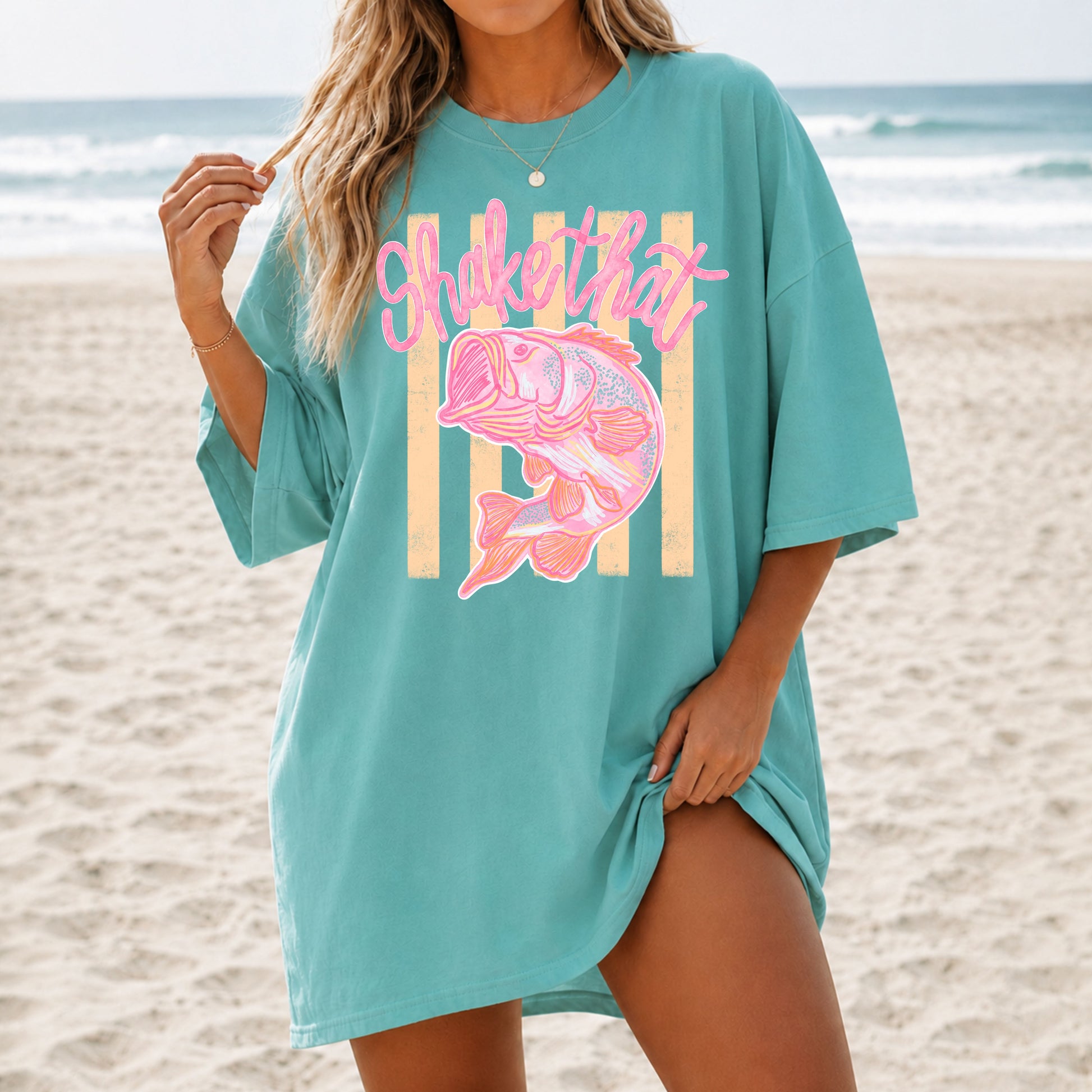 Woman wearing an oversized teal coastal graphic t-shirt featuring a pink fish illustration and the phrase "Shake That" while standing on a sandy beach with ocean waves in the background.