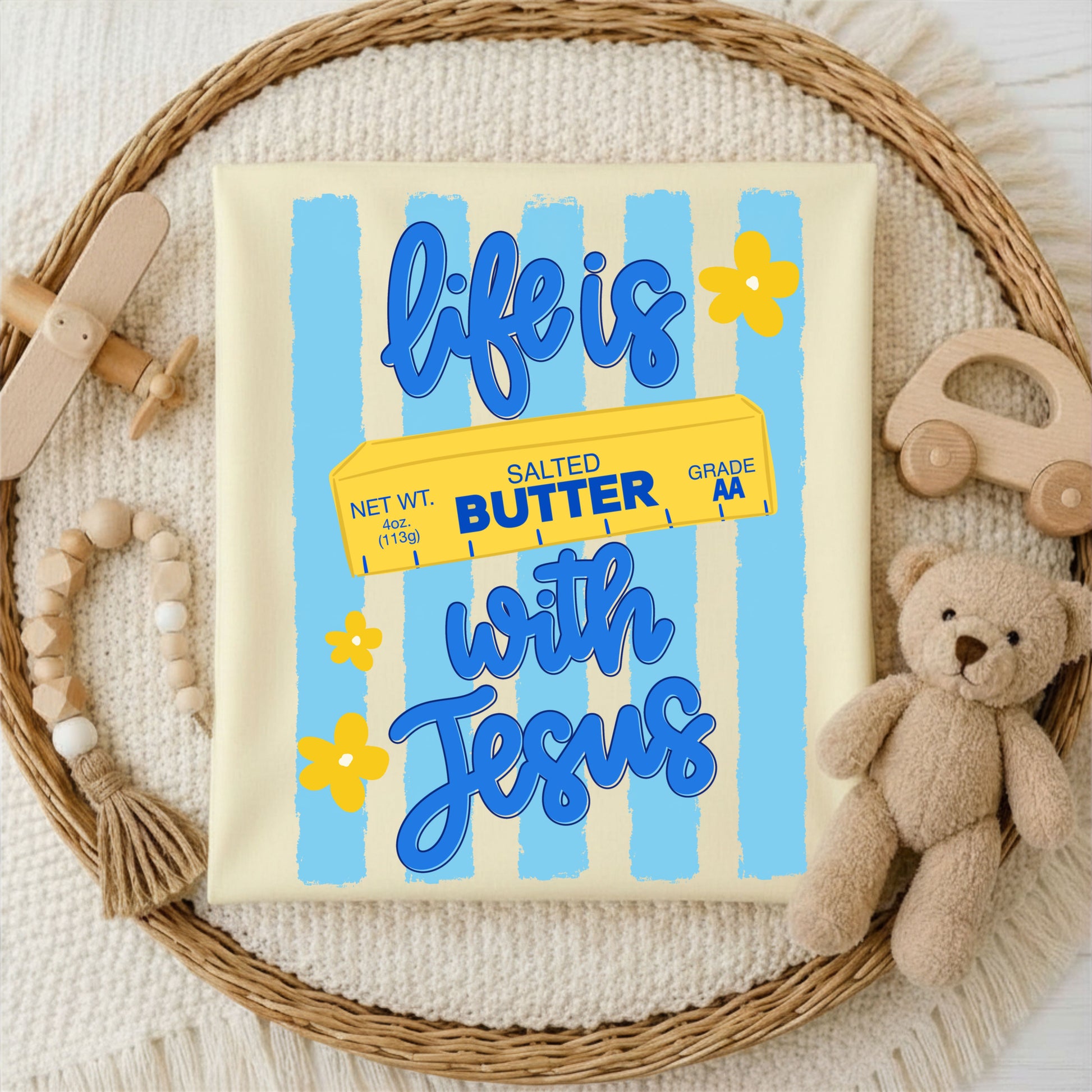 Canvas with 'life is salted butter with Jesus' text on a woven mat with toys