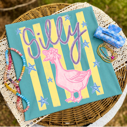 Flat lay of a teal graphic t-shirt featuring the words "Silly" above a pink goose wearing a bow with vintage cream stripes and blue stars, styled with colorful beaded bracelets, gold bracelets, and a blue gingham hair clip on a wicker table with lace fabric.