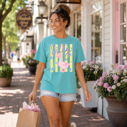 Woman wearing a teal "Silly Goose" graphic t-shirt featuring a pink bow-wearing goose with vintage stripes while walking through a charming Southern shopping district carrying a boutique shopping bag.