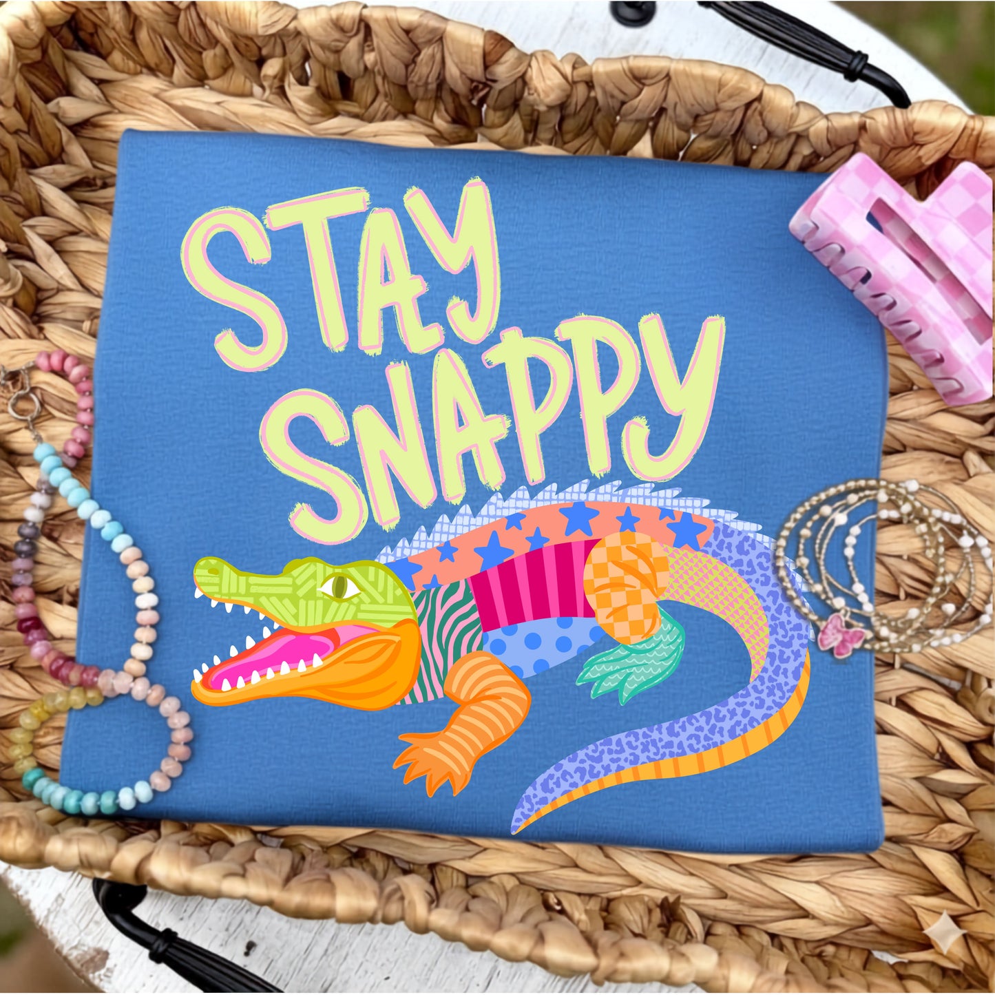 Flat lay of a royal blue graphic t-shirt featuring the phrase "Stay Snappy" above a colorful patchwork alligator, styled with pastel beaded bracelets, stacked gold bracelets, and a pink gingham hair clip on a woven tray.