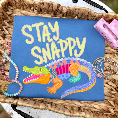 Flat lay of a royal blue graphic t-shirt featuring the phrase "Stay Snappy" above a colorful patchwork alligator, styled with pastel beaded bracelets, stacked gold bracelets, and a pink gingham hair clip on a woven tray.