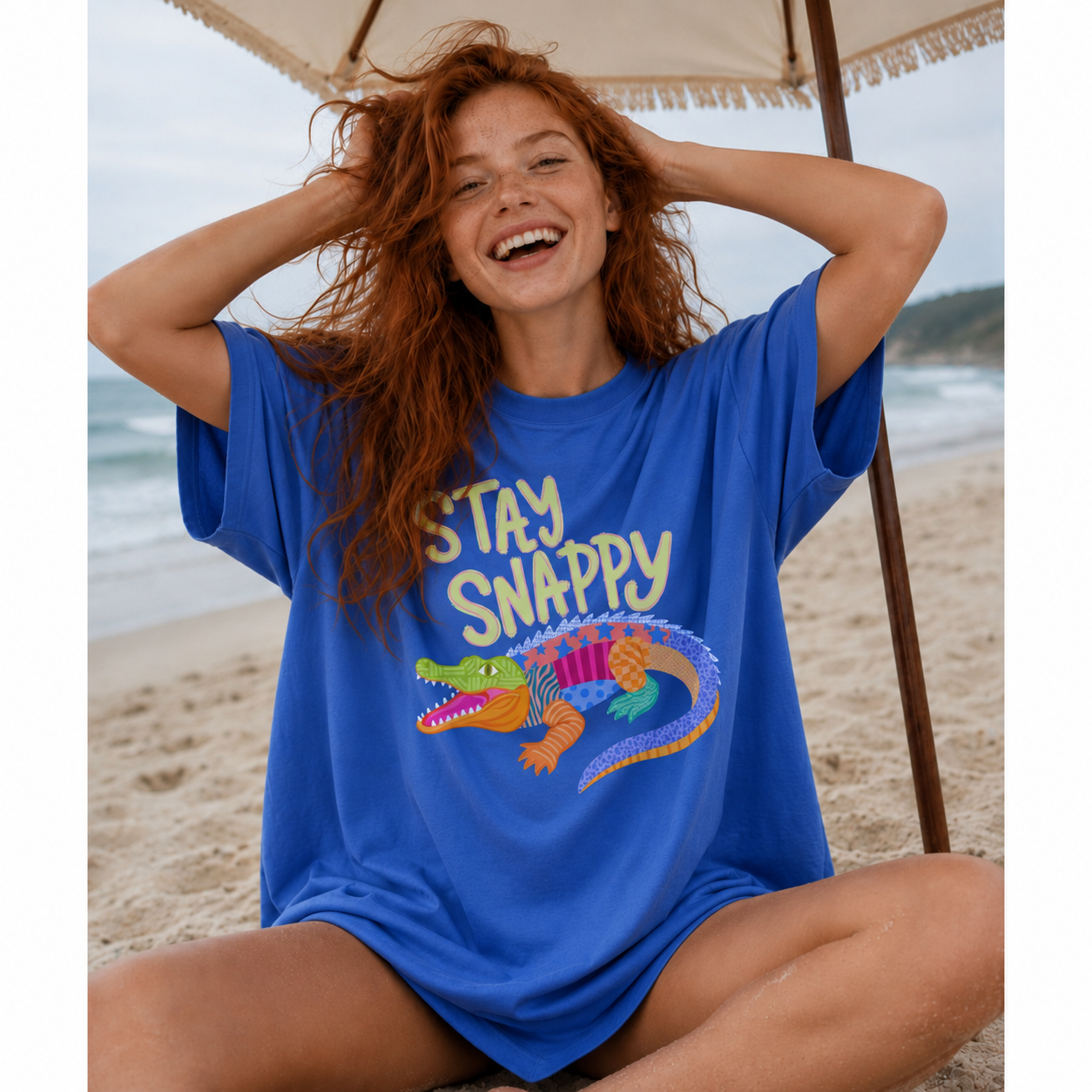 Woman wearing an oversized royal blue "Stay Snappy" graphic t-shirt featuring a colorful patchwork alligator while relaxing under a beach umbrella on the sand.