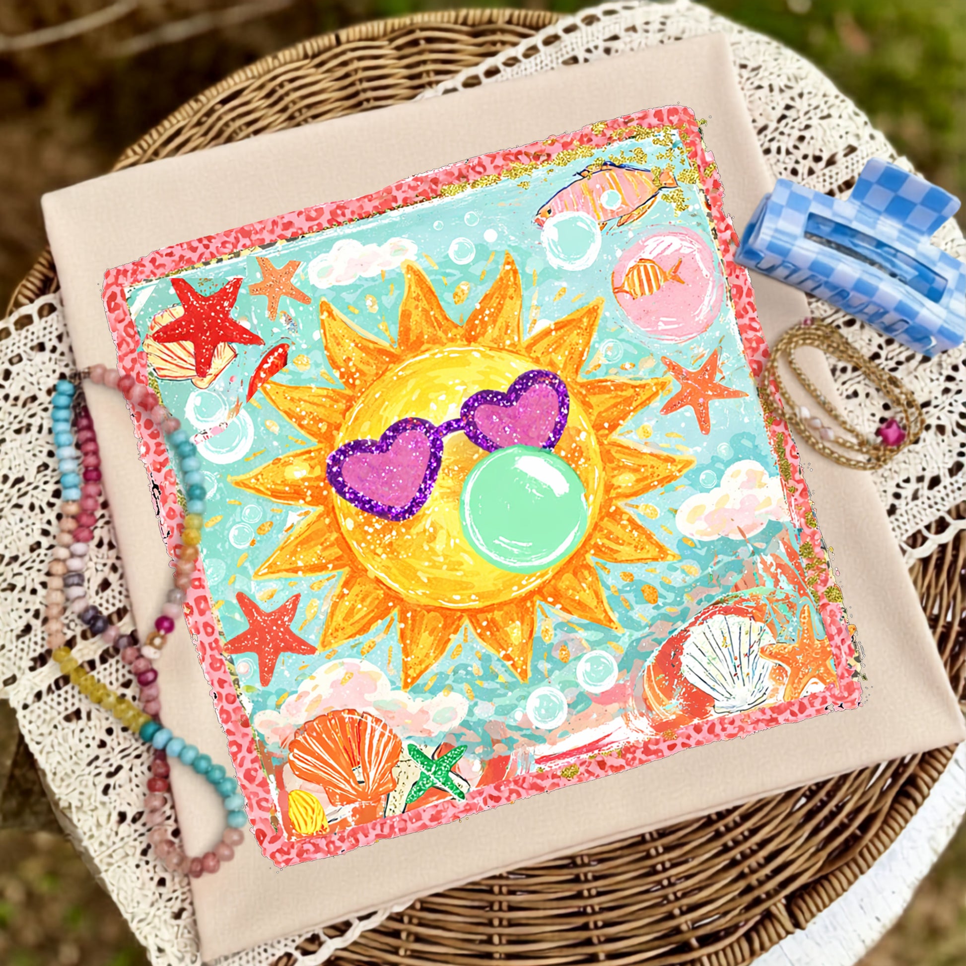Flat lay of a cream graphic t-shirt featuring a smiling sun wearing purple heart-shaped sunglasses and blowing a bubblegum bubble, surrounded by seashells, starfish, bubbles, and colorful coastal artwork, styled with pastel beaded bracelets, stacked gold bracelets, and a blue gingham hair clip on a wicker table with lace fabric.