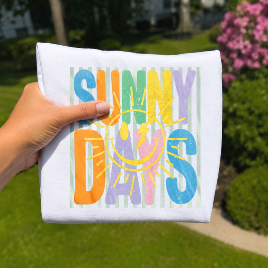 Hand holding a folded white graphic t-shirt featuring colorful "Sunny Days" lettering with a retro smiley sun and pastel striped background against a sunny outdoor garden backdrop.
