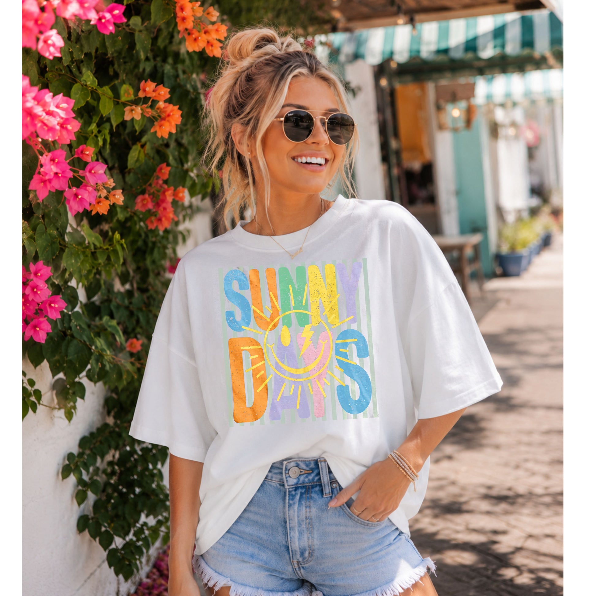 Woman wearing a white Sunny Days graphic t-shirt featuring colorful retro lettering and a smiling sunshine design, styled with light wash denim shorts while standing beside blooming flowers in a charming outdoor shopping district.