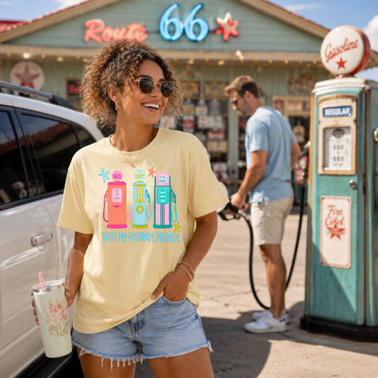 Woman wearing a butter yellow graphic t-shirt featuring colorful retro gas pumps and the phrase "That's My Husband's Problem" while smiling outside a vintage Route 66 gas station as a man fuels a vehicle in the background.