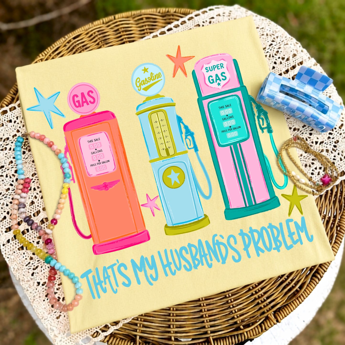 Flat lay of a butter yellow graphic t-shirt featuring three colorful retro gas pumps with the phrase "That's My Husband's Problem," styled with pastel beaded bracelets, stacked gold bracelets, and a blue gingham hair clip on a wicker table with lace fabric.