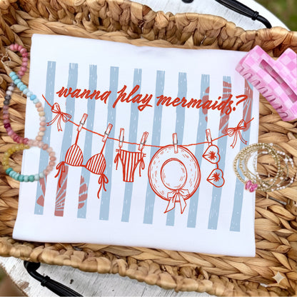 Flat lay of a white graphic t-shirt featuring the phrase "Wanna Play Mermaids?" above a whimsical clothesline with a bikini, swim bottoms, sunhat, and sunglasses over soft blue coastal stripes, styled with pastel beaded bracelets, stacked gold bracelets, and a pink gingham hair clip on a woven tray.