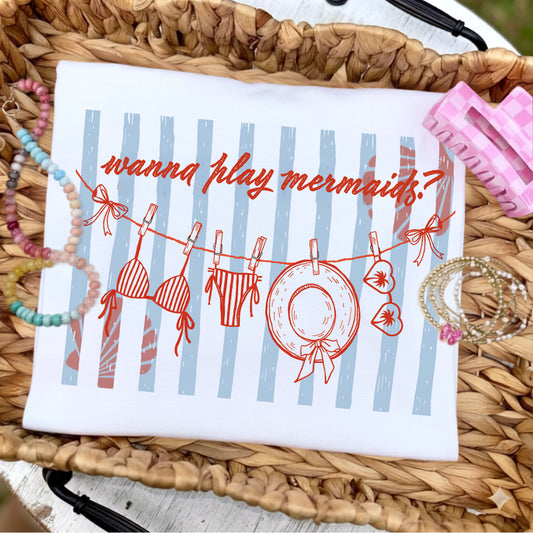 Flat lay of a white graphic t-shirt featuring the phrase "Wanna Play Mermaids?" above a whimsical clothesline with a bikini, swim bottoms, sunhat, and sunglasses over soft blue coastal stripes, styled with pastel beaded bracelets, stacked gold bracelets, and a pink gingham hair clip on a woven tray.