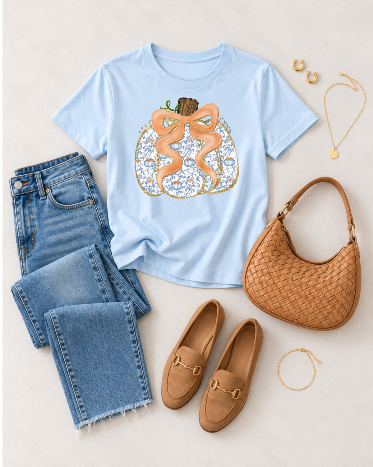 Chambray blue pumpkin bow graphic tee styled in a flat lay with jeans, tan loafers, woven bag, and gold jewelry