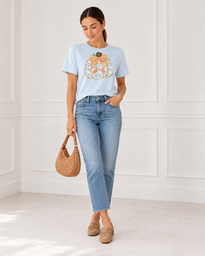 Woman wearing a chambray blue pumpkin bow graphic tee with ankle jeans, tan loafers, and a woven handbag