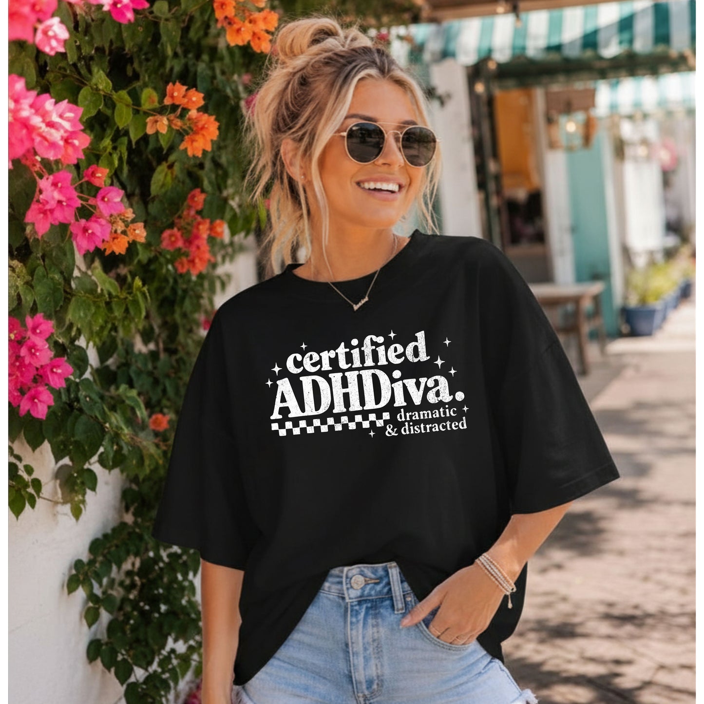 Woman standing in front of a flower covered wall while wearing a black t-shirt that says certified adhdiva.