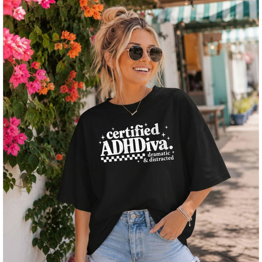 Woman standing in front of a flower covered wall while wearing a black t-shirt that says certified adhdiva.